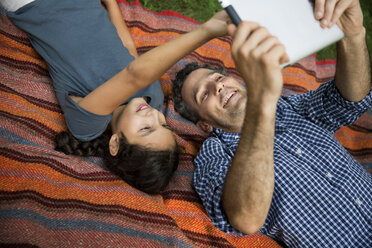 Happy father and daughter looking at tablet computer while lying on blanket in yard - CAVF40365