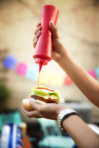 Ausgeschnittenes Bild einer Frau, die bei einer Gartenparty Ketchup auf einen Hamburger gießt, lizenzfreies Stockfoto