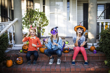 Glückliche Geschwister mit Requisiten, die mit dekorierten Kürbissen auf den Stufen sitzen, während Halloween - CAVF40543