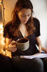Ernste Frau hält Kaffeetasse beim Lesen eines Buches zu Hause - CAVF40549