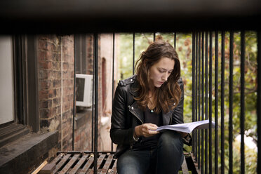 Serious woman reading book while sitting at fire escape - CAVF40557