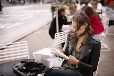 Frau in Jacke liest ein Buch, während sie in einem Straßencafé sitzt - CAVF40562