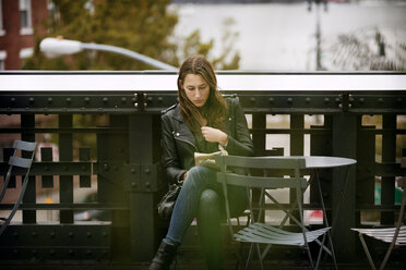 Eine Frau liest ein Buch, während sie an einem Tisch auf einer Brücke in der Stadt sitzt - CAVF40567