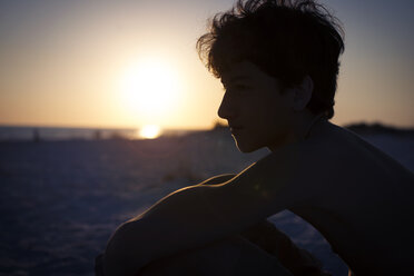 Thoughtful teenager looking away while sitting at beach during sunset - CAVF40634