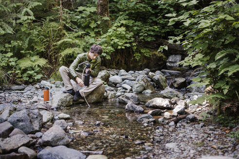 Wanderer, der auf einem Felsen am Seeufer im Wald sitzt und seine Wasserflasche auffüllt - CAVF40675