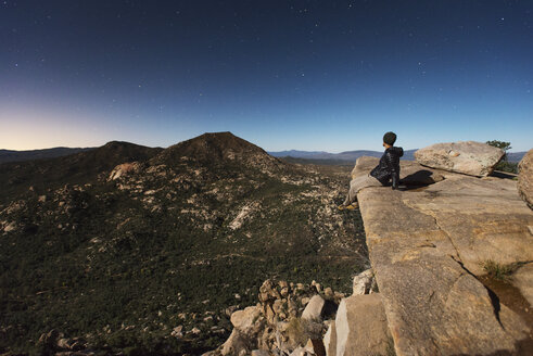 Wanderer sitzt auf einem Felsen und betrachtet die Berge gegen den Himmel in der Abenddämmerung - CAVF40703