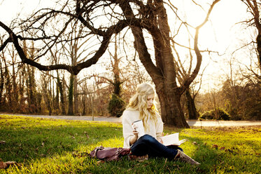 Frau liest ein Buch und hält einen Einwegbecher, während sie auf einer Wiese im Park sitzt - CAVF40736