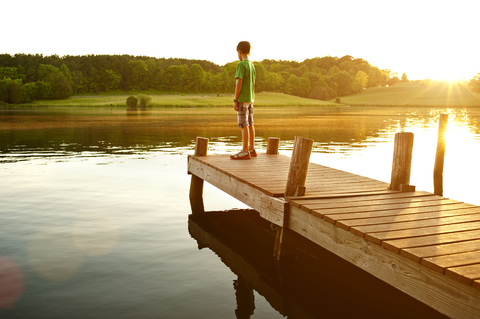 Junge steht an einem sonnigen Tag auf einem Steg über einem See, lizenzfreies Stockfoto