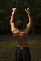 Rear view of teenager exercising on gymnastic rings on grassy field at park - CAVF40777