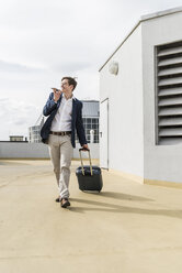 Smiling businessman with rolling suitcase using smartphone at parking garage - UUF13418