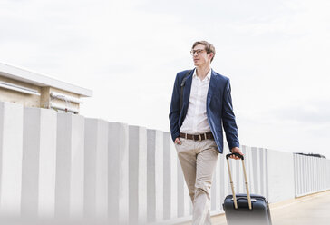 Confident businessman with rolling suitcase walking at parking garage - UUF13420
