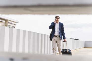 Smiling businessman with rolling suitcase using smartphone at parking garage - UUF13423