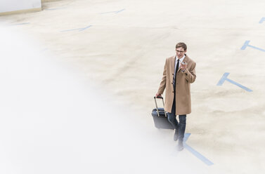 Businessman with rolling suitcase and smartphone walking at parking garage - UUF13438
