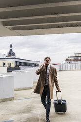 Businessman with rolling suitcase and smartphone walking at parking garage - UUF13442