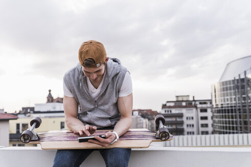 Lächelnder Mann sitzt in der Stadt mit Skateboard und benutzt ein Mobiltelefon - UUF13456