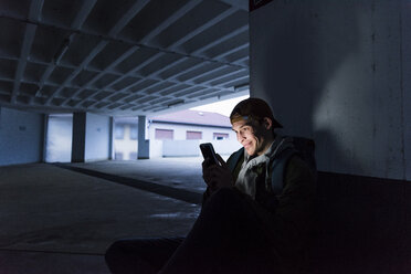 Smiling man looking at shining smartphone in parking garage - UUF13469