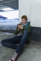 Smiling man sitting on the ground looking at smartphone in parking garage - UUF13470