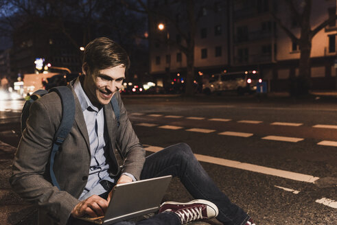 Lächelnder Geschäftsmann mit Laptop auf einer nächtlichen Straße in der Stadt - UUF13473