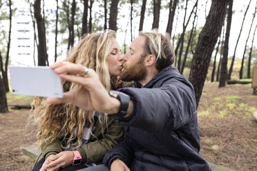 Romantisches Paar, das sich küsst, während es ein Selfie mit dem Smartphone im Wald macht - CAVF40822