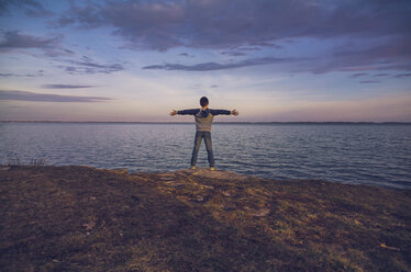 Rear view of boy with arms outstretched standing against sea and cloudy sky during sunset - CAVF40911