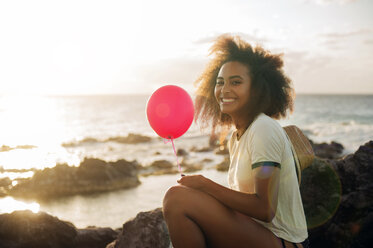 Glückliches Teenager-Mädchen, das einen Ballon hält, während es am felsigen Ufer sitzt - CAVF41308