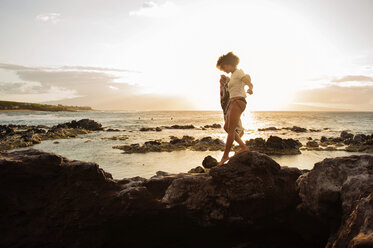 Teenager walking on top of rocks by sea against sky during sunset - CAVF41309