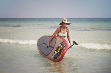 Woman carrying paddleboard while walking in sea against clear sky - CAVF41397