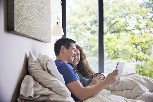 Glückliches Paar, das sich unterhält und einen Tablet-Computer benutzt, während es zu Hause auf dem Bett liegt - CAVF41557