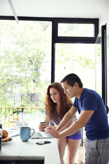 Ehepaar schaut auf den Laptop, während sie an der Küchentheke am Fenster Kaffee trinken - CAVF41567