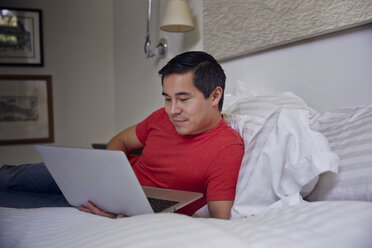 Mann benutzt einen Laptop, während er zu Hause auf dem Bett liegt - CAVF41577