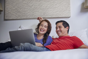 Fröhliches Paar schaut auf einen Laptop, während es zu Hause auf dem Bett liegt - CAVF41578