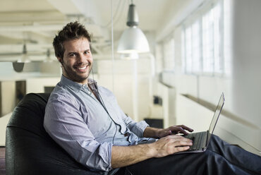 Portrait of smiling businessman using laptop while sitting on bean bag in office - MASF04678