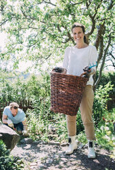 Porträt einer glücklichen Frau, die einen Weidenkorb trägt, mit einem Mann bei der Gartenarbeit im Hintergrund im Garten - MASF04688