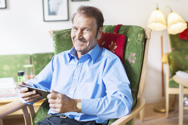 Porträt eines glücklichen älteren Mannes, der ein digitales Tablet in der Hand hält und auf einem Sessel im Pflegeheim sitzt - MASF04709