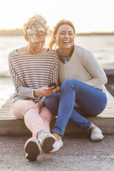 Portrait of happy woman sitting with female friend using mobile phone on pier - MASF04717