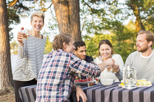 Porträt einer glücklichen Frau, die ein Bierglas hält, während sie bei Freunden steht, die am Picknicktisch zu Mittag essen - MASF04719