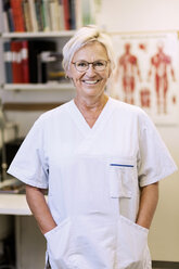 Portrait of happy senior orthopedic surgeon standing with hands in pockets in clinic - MASF04725