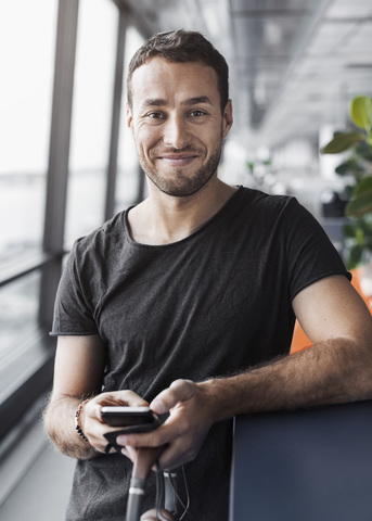 Porträt eines lächelnden Geschäftsmannes, der ein Mobiltelefon hält, während er sich an eine Kabine im Büro lehnt, lizenzfreies Stockfoto