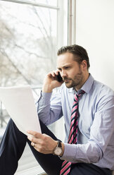 Serious mid adult businessman reading document while on call at office - MASF04741