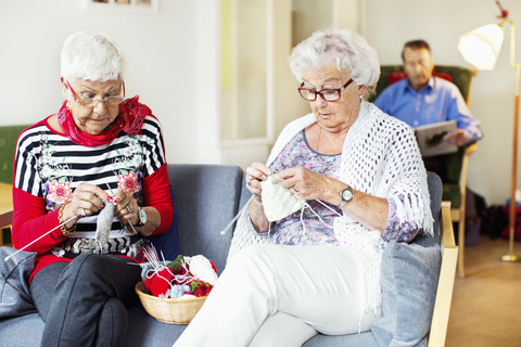 Eine ältere Frau strickt, während ein Mann im Hintergrund ein Buch liest, lizenzfreies Stockfoto