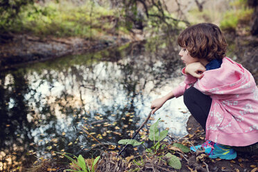 Side view of girl holding stick while sitting by stream in forest - CAVF41683