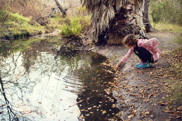 Side view of girl holding stick while crouching by stream - CAVF41684