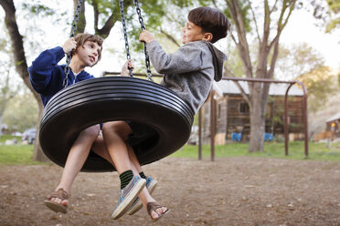 Side view of siblings sitting on tire swing at park - CAVF41698