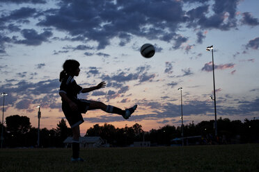 Side view of girl kicking soccer ball at field against sky during sunset - CAVF41717
