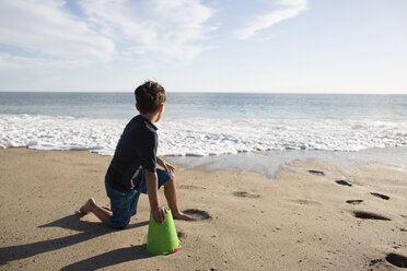 Junge schaut auf das Meer, während er mit einem Eimer am Strand in der Sonne sitzt - CAVF41739