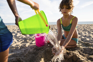 Junge gießt Wasser auf die Hände seiner Schwester beim Bau einer Sandburg am Strand - CAVF41743