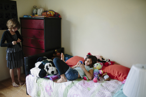 Mädchen, das sich anzieht, während seine Schwester auf dem Bett zu Hause ein Mobiltelefon benutzt, lizenzfreies Stockfoto