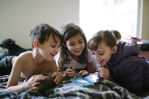Glückliche Geschwister, die zu Hause auf dem Bett ihr Handy betrachten, lizenzfreies Stockfoto
