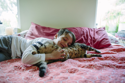 Lächelnder Junge schläft mit Katze auf dem Bett zu Hause, lizenzfreies Stockfoto