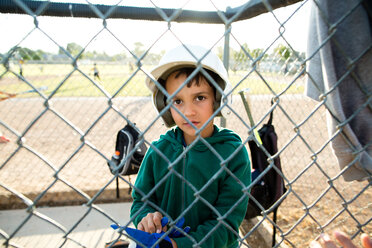 Porträt eines Jungen mit Baseballhelm, der im Unterstand sitzt, gesehen durch einen Maschendrahtzaun - CAVF41837
